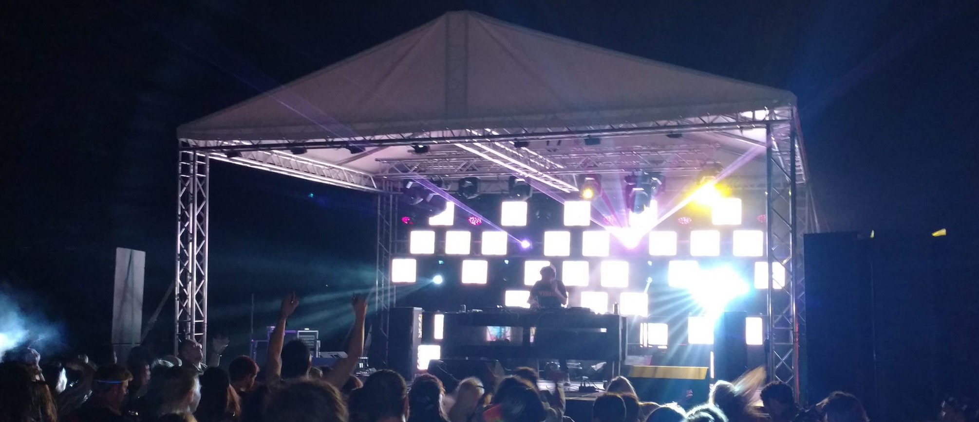 Nighttime festival stage structure tent with aluminum trussing, LED lighting, DJ equipment, and crowd at a music event